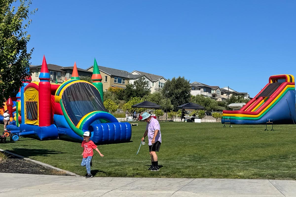 people at large grassy park enjoying playing with jumpers/bounce houses and interactive games and slides